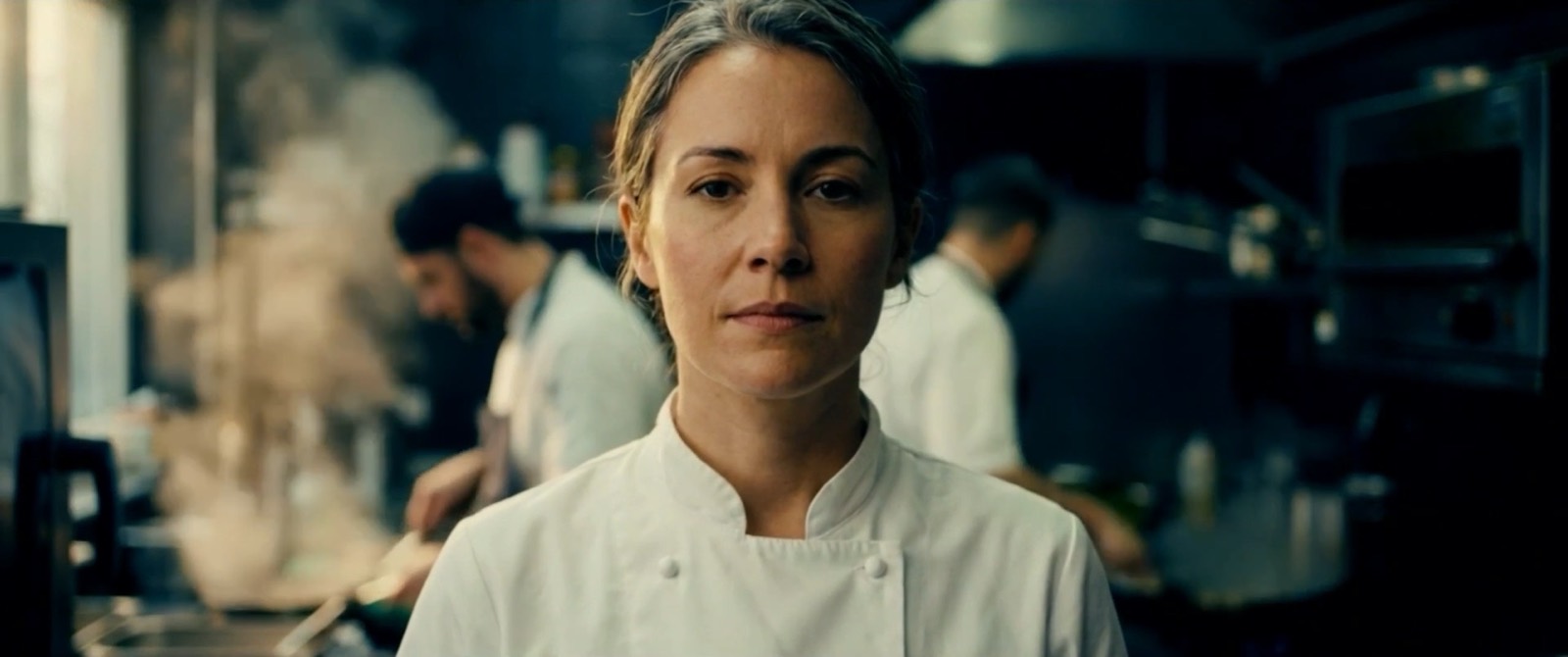 A chef in whites looking straight down the lens, her kitchen at work and out of focus behind her. A frame from the series trailer.